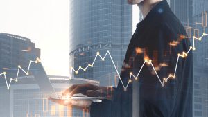 Professional standing in front of skyscrapers holding a laptop and analyzing market performance.