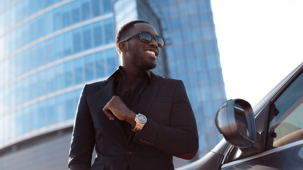 Man in front of skyscraper with nice car, watch, suit and sunglasses.