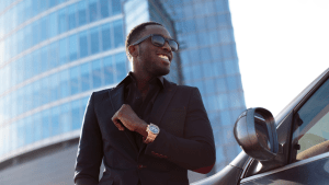 Man in front of skyscraper with nice car, watch, suit and sunglasses.