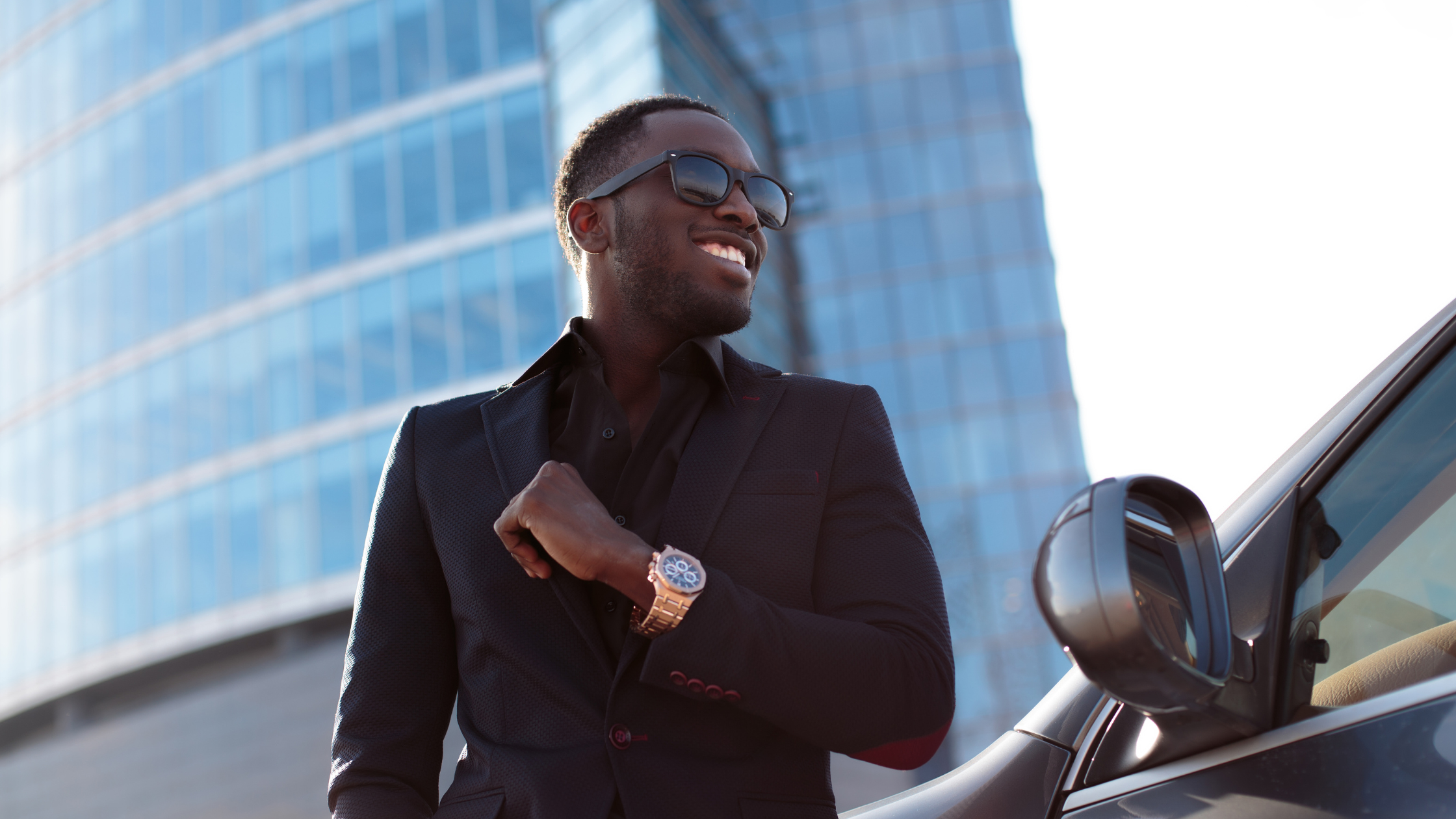 Man in front of skyscraper with nice car, watch, suit and sunglasses.