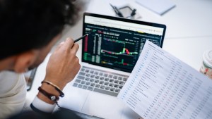 Two people reviewing stock market on computer and printed results.