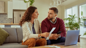 A couple sitting on the couch with a laptop on the coffee table. The woman is holding documents. They are in conversation.