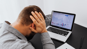 Man looks at laptop showing market downturn. Head in hands.