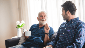 Older gentleman and son having a conversation on a couch in front of a window with white blinds. Older gentleman is holding a coffee cup in his right hand and gesticulating with left hand.
