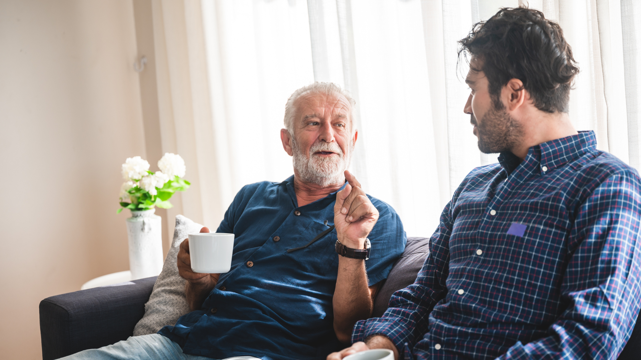 Older gentleman and son having a conversation on a couch in front of a window with white blinds. Older gentleman is holding a coffee cup in his right hand and gesticulating with left hand.