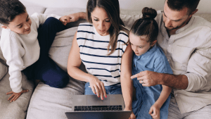 Family sitting on couch reviewing something on laptop. Mom is holding laptop, Dad is pointing to screen, daughter is sitting inbetween them and son is on the side.