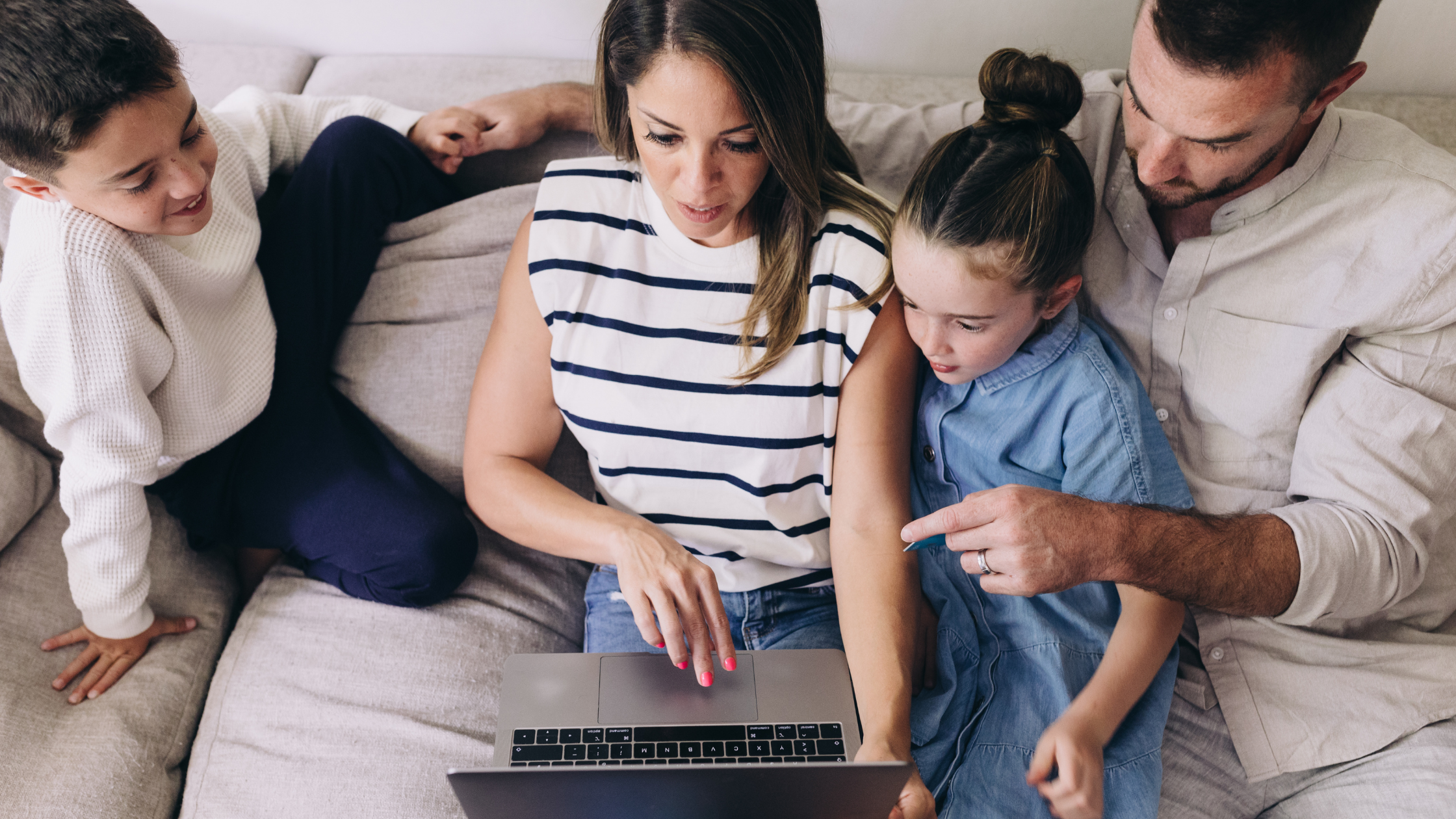 Family sitting on couch reviewing something on laptop. Mom is holding laptop, Dad is pointing to screen, daughter is sitting inbetween them and son is on the side.